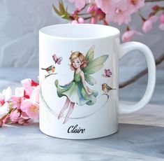 a white coffee mug with a green fairy sitting on top of it next to pink flowers