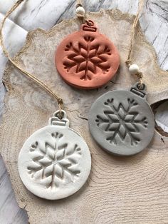 three clay ornaments are sitting on a piece of wood, one has a snowflake ornament