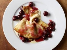 a white plate topped with crepes covered in sauce and cherries on top of a wooden table