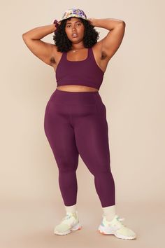 a woman in a purple sports bra top and matching leggings stands with her hands on her head