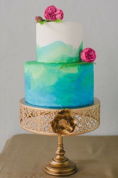 a three tiered cake with blue and green frosting on a gold plated stand