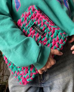 a person wearing a green sweater holding a pink and blue purse in their hands while standing next to a potted plant