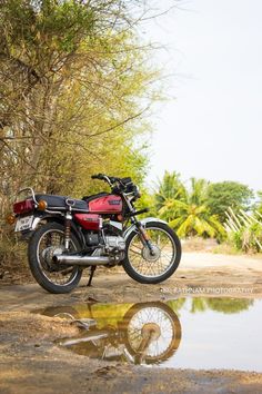 a red motorcycle parked on the side of a dirt road next to trees and water