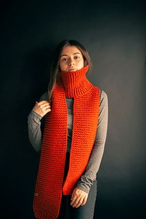 a woman wearing an orange knitted scarf and grey shirt standing in front of a black wall