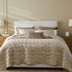 a bed with beige comforter and pillows in a bedroom next to a lamp on a dresser