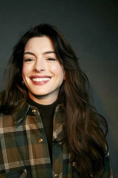 a woman with long hair smiling and wearing a green plaid jacket, black turtle neck sweater and gold hoop earrings