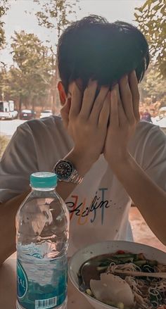 a man covering his face while sitting at a table with food and water in front of him