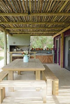 a wooden table sitting under a pergolated roof next to a fire place with flowers in a vase