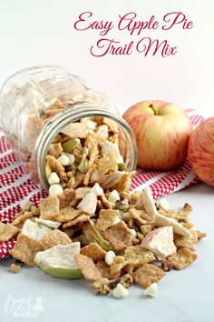 an apple pie trail mix in a glass jar next to two apples on a red and white checkered table cloth