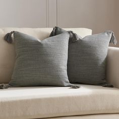 two gray pillows sitting on top of a white couch next to each other with tassels