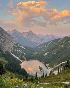 a lake in the middle of mountains surrounded by grass and flowers with clouds above it