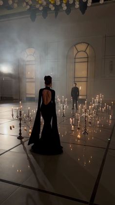 the back of a woman in a black dress walking through a room filled with candles