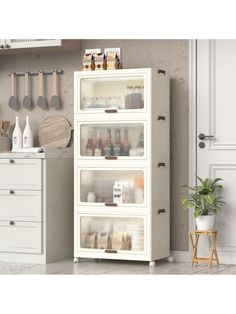 a white cabinet filled with lots of items next to a potted plant and wooden spoons