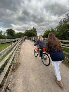 two people riding bikes on a dirt road near a fence and grassy area with trees in the background