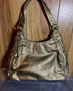 A vintage uber adorable Coach gold metallic purse. Can be used as a shoulder or a handbag. Coach Madison Maggie gold metallic patent leather satchel bag,style 14331. It is about 8.1/4"H  14"L 6"D 9 1/2" drop. Double handles, front has Coach logo and stagecoach on the top center, 3 Coach hangtags of various sizes are attached to one side, pleats, and is very pliable, 3 compartments, lining is a beautiful light blue, like satin feel. The center area has a top zipper with a patent leather matching Metallic Purse, Coach Logo, Leather Satchel Bag, Coach Purse, Bag Style, Leather Pulls, Vintage Coach, Satchel Bag, Coach Purses