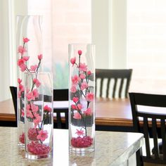 two clear vases with pink flowers in them sitting on a marble counter top next to a dining room table