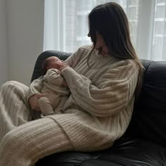 a woman sitting on a couch holding a baby wrapped in a blanket and looking out the window