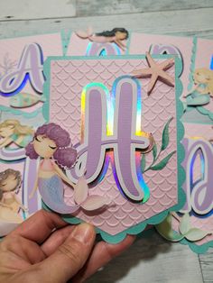 a hand holding up a card with mermaid stickers on it's side and the letter m in the middle