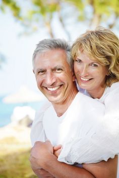 an older couple hugging each other in front of a tree and the ocean behind them