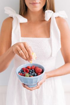 a woman in white dress holding a bowl of berries and oranges with lemon wedge