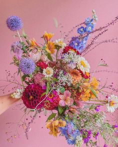 a person holding a bouquet of flowers in their hand against a pink background with grass and wildflowers