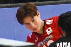 a young man in a red shirt is playing ping pong with another person behind him