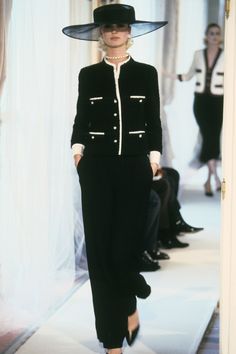 a woman walking down a runway wearing a black suit and hat with pearls on it