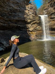 a woman sitting on top of a rock next to a body of water with a waterfall in the background