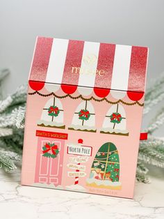 a pink and white box with christmas decorations on the front, sitting next to a tree