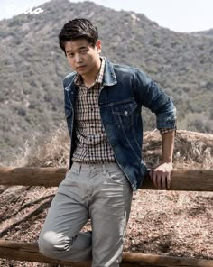 a young man leaning on a wooden fence in front of a mountainous area with trees and bushes