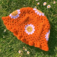 an orange crocheted hat with white flowers on it sitting in the grass next to daisies