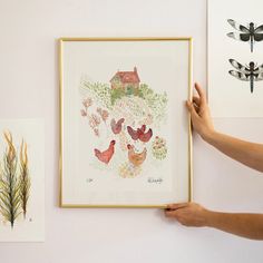 a person holding up a framed painting next to two dragonflys on the wall