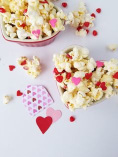 two heart shaped bowls filled with popcorn on top of a white table next to hearts and confetti