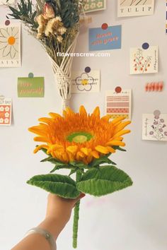 a hand holding a fake sunflower in front of a wall with pictures on it