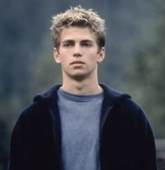 the young man is wearing a blue shirt and black cardigan jacket while standing in front of some trees