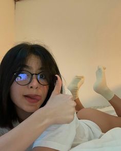 a woman laying on top of a bed with her hand up to her face and wearing glasses