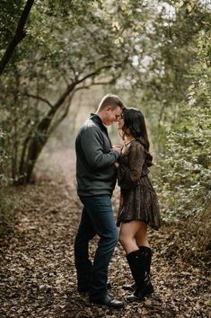 a man and woman standing in the woods with leaves on the ground, looking into each other's eyes