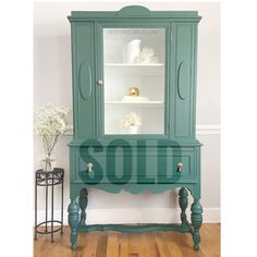 a green china cabinet sitting on top of a hard wood floor next to a vase with flowers