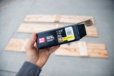 a person holding up a black box with yellow stickers on it and some wood planks in the background