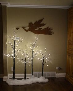 three trees with fairy lights on them are in front of a wall that has an angel cutout