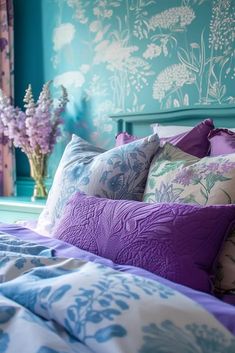 a bed with purple and blue pillows on top of it next to a vase filled with flowers