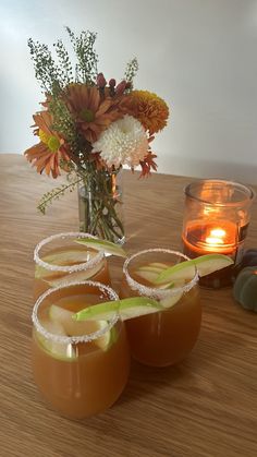 three glasses filled with drinks sitting on top of a wooden table next to a vase full of flowers