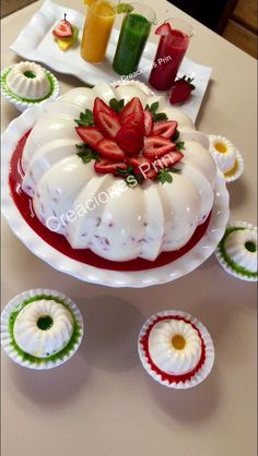 a white cake with strawberries on top and cupcakes next to it sitting on a table