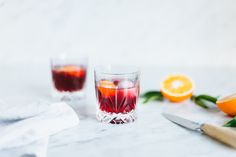 two glasses filled with red liquid next to sliced oranges and a knife on a table