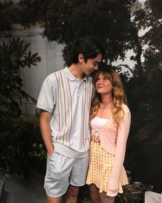 a young man and woman standing next to each other in front of a wall with trees