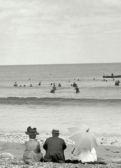 two people sitting on the beach under umbrellas looking out at the ocean and swimmers