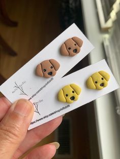 two small yellow and brown dog shaped buttons in their packaging, one being held up by someone's hand