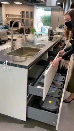 a woman standing next to a sink in a kitchen with an open drawer underneath it