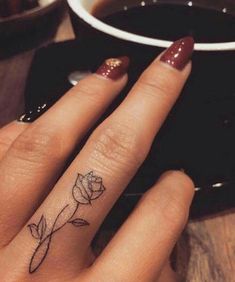 a woman's hand with a small rose tattoo on her left ring finger next to a cup of coffee