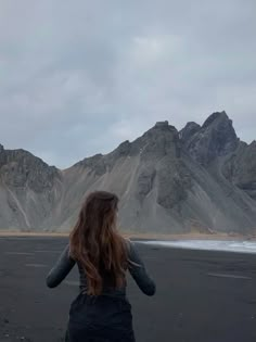 a woman standing on top of a black beach under a cloudy sky with mountains in the background